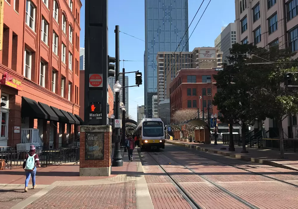 A light rail train approaching in a downtown street lined with brick buildings and skyscrapers in the background.